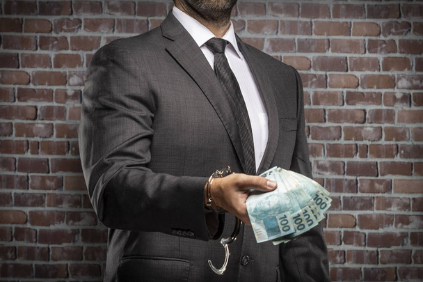 Brazilian man holding bills of money with a handcuff in a jail. concept of corruption, corrupt politicians, illegal businesses. brick background. - Fotó, kép