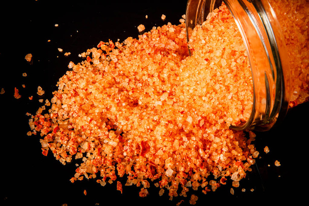 Closeup velké oranžové přírodní organická mořská sůl vylit z malé skleničky na pozadí černé zrcadlo - Fotografie, Obrázek