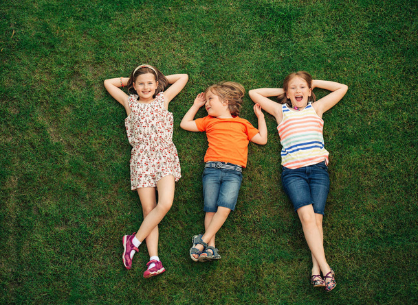 Szczęśliwe dzieci, zabawy na świeżym powietrzu. Dzieci gry w lato park. Mały chłopiec i dwie dziewczyny leżącej na zielonej trawie świeże - Zdjęcie, obraz