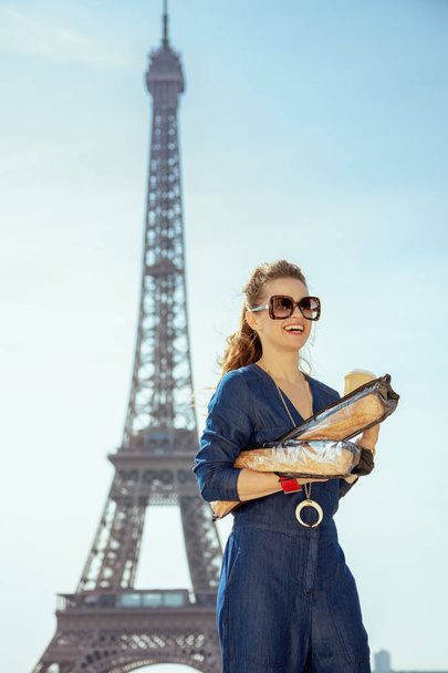 happy stylish woman in blue jeans overall with coffee cup and two baguettes looking into the distance in Paris, France. - Фото, изображение