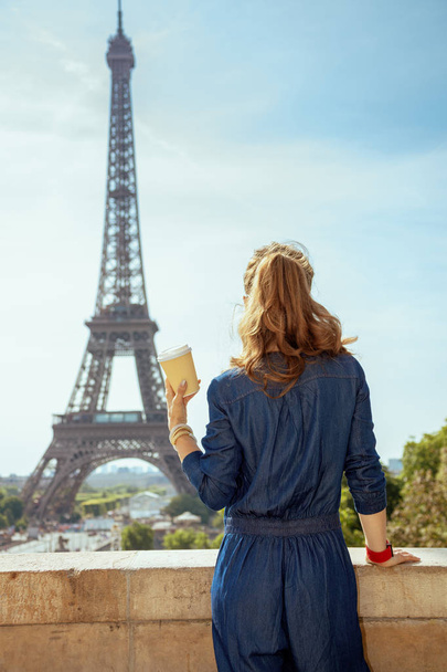Seen from behind trendy tourist woman in blue jeans overall with coffee cup in Paris, France. - Фото, изображение