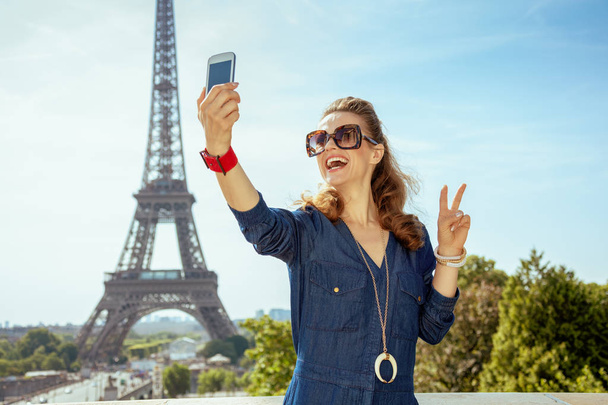 happy trendy woman in blue jeans overall taking selfie with cellphone and showing victory at Trocadero overlooking Eiffel tower in Paris, France. - Zdjęcie, obraz