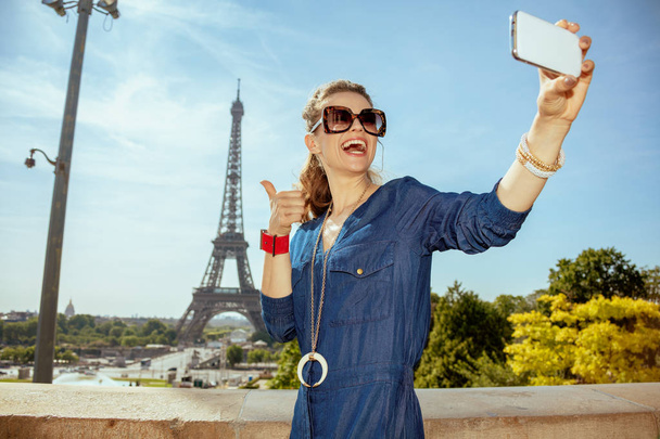 happy trendy woman in blue jeans overall showing thumbs up and taking selfie with smartphone in Paris, France. - Fotoğraf, Görsel