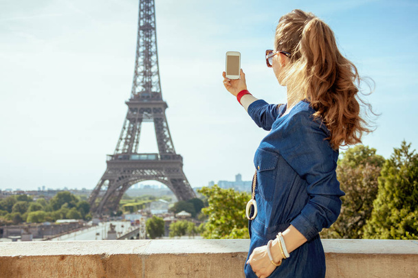 Seen from behind trendy woman in blue jeans overall taking photo with smartphone against Eiffel tower in Paris, France. - Zdjęcie, obraz