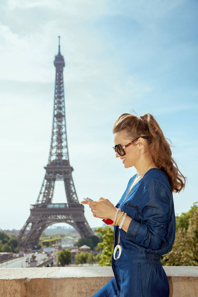 ευτυχισμένη γυναίκα κομψό στο μπλε τζιν συνολικά στο μπροστινό μέρος του Πύργου του Άιφελ στο Παρίσι, Γαλλία βλέποντας βίντεο στο διαδίκτυο μέσω κινητού τηλεφώνου. - Φωτογραφία, εικόνα