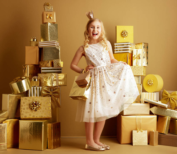Ganzkörperporträt einer glücklichen, modernen kleinen Prinzessin in beigefarbenem Fit-and-Flare-Kleid und einer kleinen Krone auf dem Kopf mit goldenen Einkaufstaschen zwischen zwei Stapeln goldener Geschenke vor einer schlichten Wand. - Foto, Bild