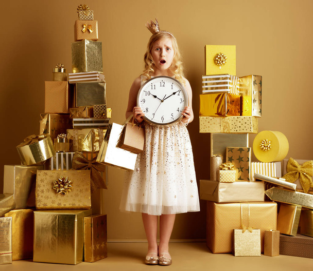 Full length portrait of shocked elegant girl in beige fit and flare dress and a little crown on head with gold shopping bags and round clock among 2 piles of golden gifts in front of a plain wall. - Valokuva, kuva