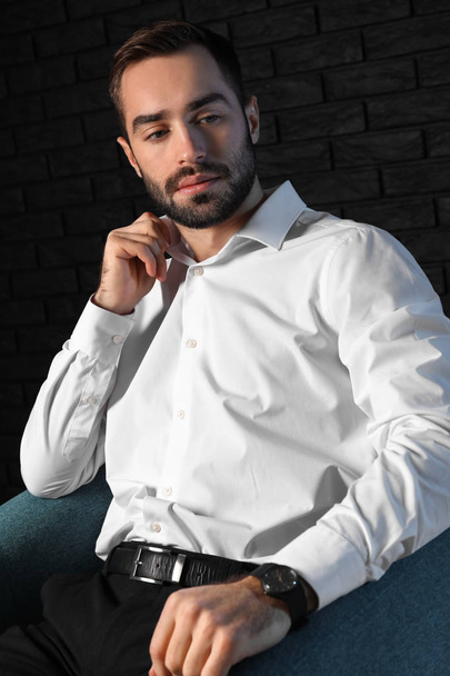 Handsome fashionable man sitting in armchair against dark background - Foto, Imagem