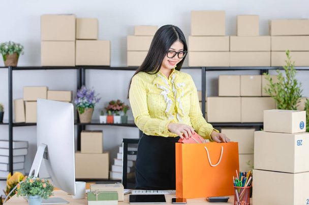 Young attractive Asian businesswoman, long black hair, wearing glasses, yellow blouse, prepare gift boxes for customer, put in orange shopping bag, in startup office background full of boxes - Φωτογραφία, εικόνα