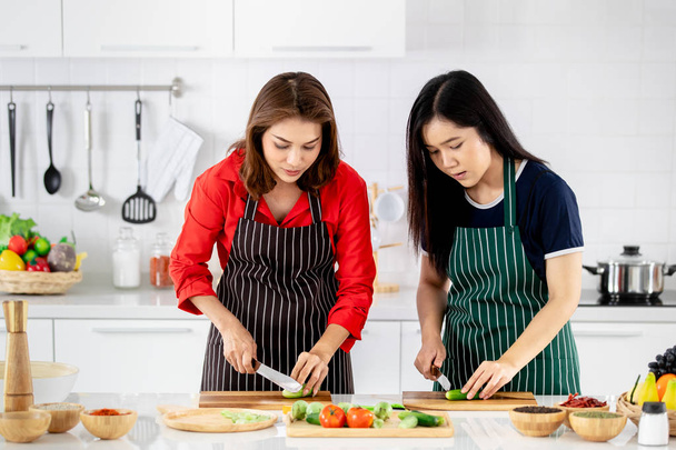 Gyönyörű ázsiai nő a piros inget és fekete kötény tanítása, a lánya fehér tiszta modern konyha főzés a zöldségek felkészülni. - Fotó, kép