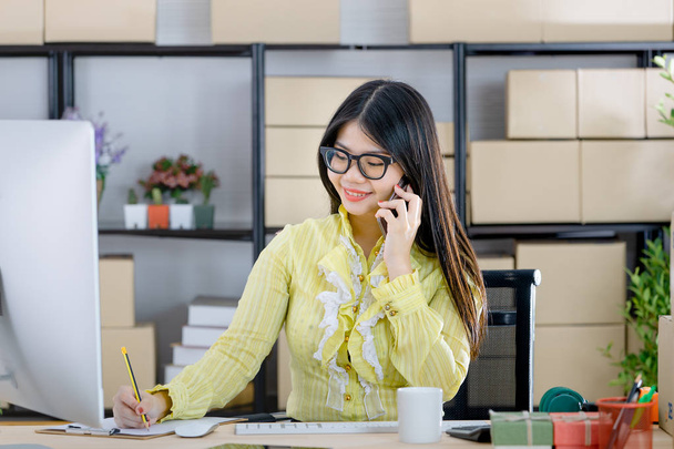 Young attractive Asian businesswoman, long black hair, wearing glasses, yellow blouse, talking on smartphone with customer, taking note, look at computer in startup office background full of boxes - Photo, Image