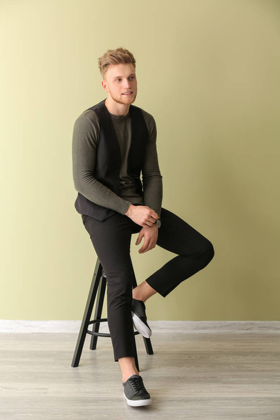 Fashionable young man sitting on stool against color wall - Foto, afbeelding