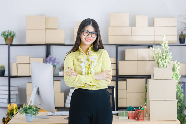 Молодая привлекательная азиатская бизнесвумен, длинные черные волосы, в очках, желтая блузка, стоя скрестив руку, уверенно улыбается в камеру с компьютером и коробками на фоне стартап-офиса
 - Фото, изображение
