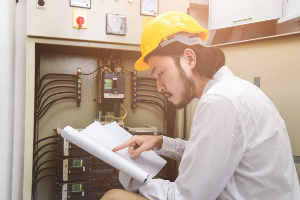 Крупный план молодого азиатского электроинженера в бежевых штанах и белой рубашке, носить желтый шлем, приседать рядом с панелью управления, рабочая инструкция в руке, следить за его повседневной проверки
 - Фото, изображение