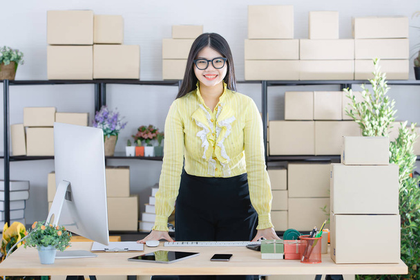 Jovem atraente mulher de negócios asiática, cabelo preto longo, usando óculos, blusa amarela, de pé, mão em cima da mesa, confiantemente sorrir para a câmera com computador e caixas no fundo do escritório de inicialização
 - Foto, Imagem