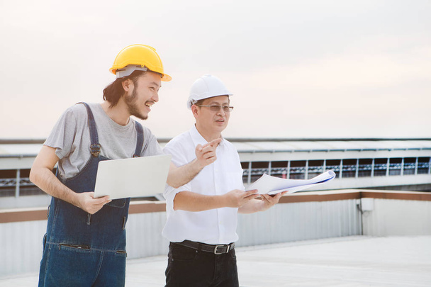 Middle age Asian factory manager and young Asian engineer in jeans jumpsuit, safety shoes, helmets, checking Lightning Rod condition on the rooftop of manufacturing building, laptop in hand - Photo, Image