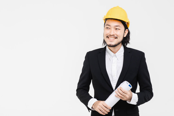 Portrait of engineer, in black suit, yellow helmet on his head, standing, he looking at the side, blueprint in his hands, white background behind him. - Photo, Image