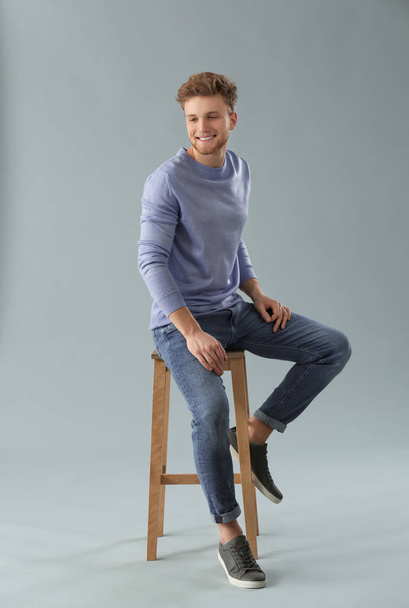 Fashionable young man sitting on stool against grey background - Foto, imagen