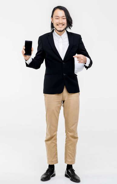 Portrait of young Asian engineer , in black suit, use cellphone, white helmet in his arm, standing, white background behind him. - Foto, Imagem