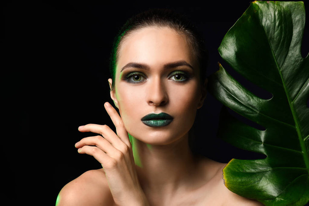 Όμορφη νεαρή γυναίκα με λαμπερό μακιγιάζ και τροπικά φύλλα σε σκούρο φόντο - Φωτογραφία, εικόνα