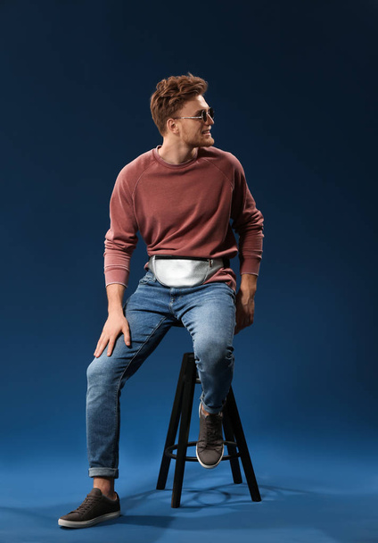 Fashionable young man sitting on stool against color background - Фото, изображение