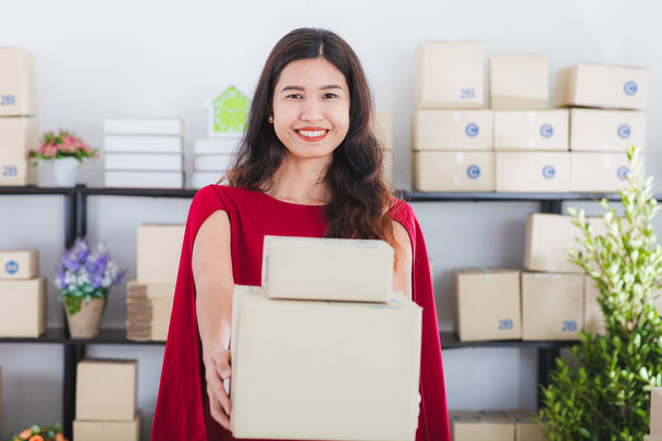 Jonge mooie, Aziatische zakenvrouw glimlachen met kuiltjes, lang zwart haar, in styling rode jurk, houd kartonnen dozen, staan voor de tafel, genieten van het werken op haar mode startup kantoor - Foto, afbeelding