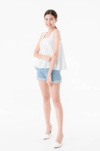Studio πορτρέτο του νεαρή όμορφη, ελκυστική γυναίκα της Ασίας σε casual καλοκαίρι ρούχα, τζιν σορτς και χαλαρά δεξαμενή κορυφή, πλατύ χαμόγελο, στέκεται, θέτοντας σε απομονωμένες άσπρο φόντο - Φωτογραφία, εικόνα