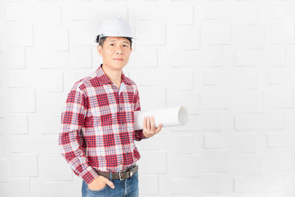 Środku wieku azjatyckich przystojnego architekta, w dżinsy i koszula w kratę, noszenie biały kask, stojący, korzystanie smartphone, rolki plany budowy pod ramieniem, na tle białej ściany - Zdjęcie, obraz