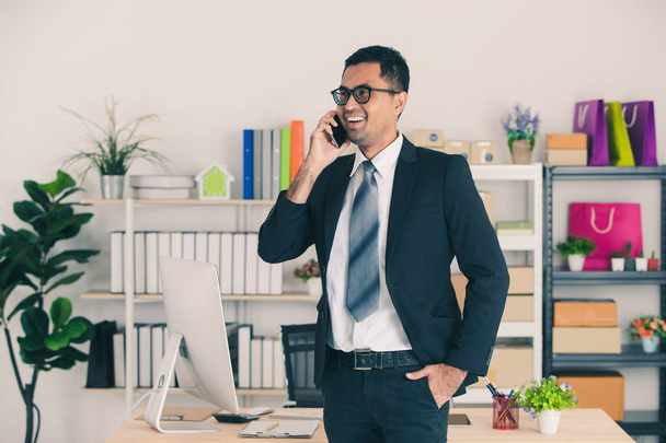 Портрет молодого, красивого, азиатского бизнесмена в очках, в деловом костюме, разговаривающего по смартфону со своим клиентом за рабочим столом со стартап-офисом
 - Фото, изображение