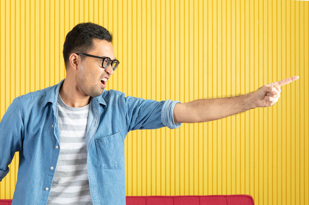 Nahaufnahme eines jungen asiatischen gutaussehenden bärtigen Mannes, der eine Brille trägt, in Jeanshemd zeigt und schreit, auf sein Team schimpft, mit gelbem Hintergrund - Foto, Bild