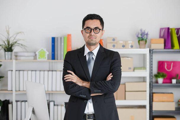 Portrét mladý, pohledný, asijské, podnikatel, nosí brýle, v obleku, stojící paže zkřížené s jistotu, na jeho pracovní stůl s spuštění office pozadí - Fotografie, Obrázek