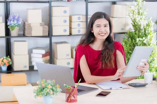 Портрет молодой красивой, привлекательной азиатской предпринимательницы в красном платье, улыбающееся лицо, сидящей на рабочем месте, проверяющей список заказов клиентов на планшете, в современном офисе стартапа
 - Фото, изображение