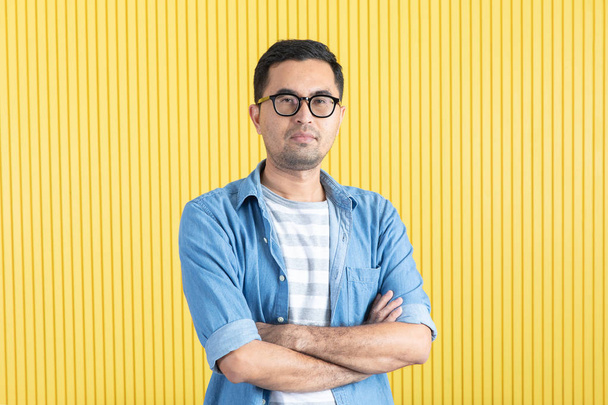Vue de face, gros plan portrait de jeune homme barbu asiatique, portant des lunettes, chemise en denim, pointant le bras croisé, main sous le menton, regarder la caméra, avec fond mural à rayures jaunes
 - Photo, image