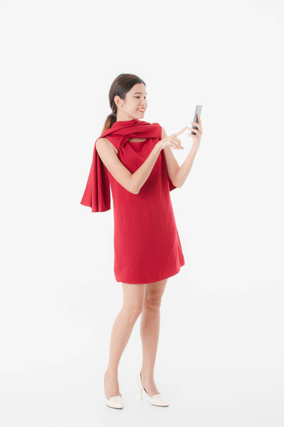 Полный рост, портрет молодой красивой, привлекательной азиатской женщины в стильном красном коротком платье, на высоком каблуке, улыбающейся, держащей в руках смартфон, посылку, сумку с покупками, на изолированном белом фоне
 - Фото, изображение