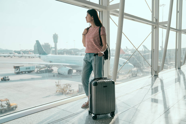 full length of travel female tourist standing with luggage watching outdoor view at airport window. chinese woman at lounge looking at airplanes while waiting at boarding gate before departure. - Photo, Image
