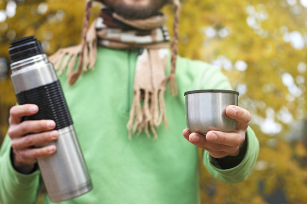 man in knitted scarf, offers a hot drink - tea or coffee from thermos to someone, front view, on the background of yellow autumn foliage - Photo, Image