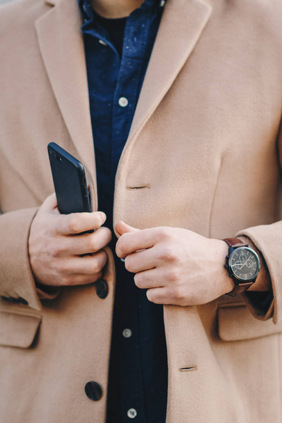 男性の手が男性のチェスター フィールド コートを携帯電話や手の時計のようなアクセサリーを押しながらボタンのディテール ショットを閉じる. - 写真・画像