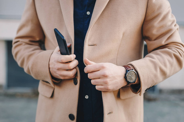 Close-up detail shot of male hands straightening men's chesterfield overcoat, while carrying accessories like a mobile phone and a hand watch. - Photo, image