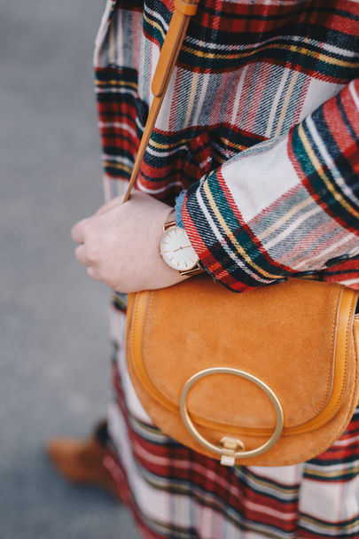 Nahaufnahme von modischen Details, stilvolle junge Frau in einem Mantel mit Schottenmuster und Armbanduhr in der Hand einer schicken Kameltasche. - Foto, Bild