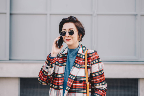 Portrait of pretty young woman having a mobile phone conversation, posing in fancy clothes in front of a building. Horizontal image. - Foto, Imagen