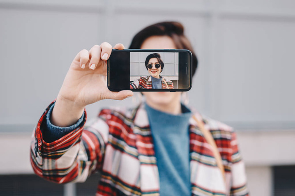 Close-up portrait of pretty and modern young blogger taking a selfie with her mobile phone, wearing fashionable clothes and posing for friends on social media. Focus on smartphone screen. - 写真・画像