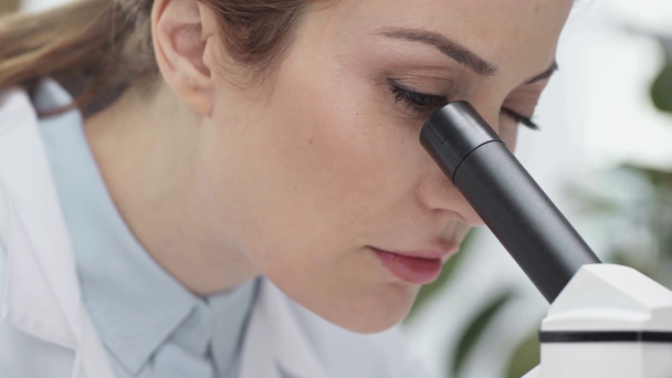 koncentruje się kobieta naukowiec w biały płaszcz patrząc przez Mikroskop w laboratorium - Materiał filmowy, wideo