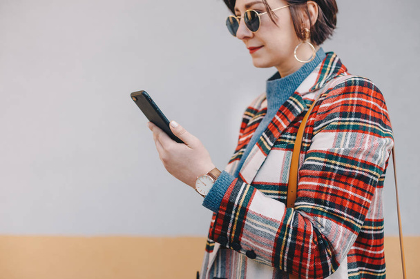 Close up, fashionable young woman using a phone, wearing plaid women's coat, sunglasses, a watch and a shoulder bag, in front of multicolored background. Space for copy, text, advertising. - Foto, Imagen