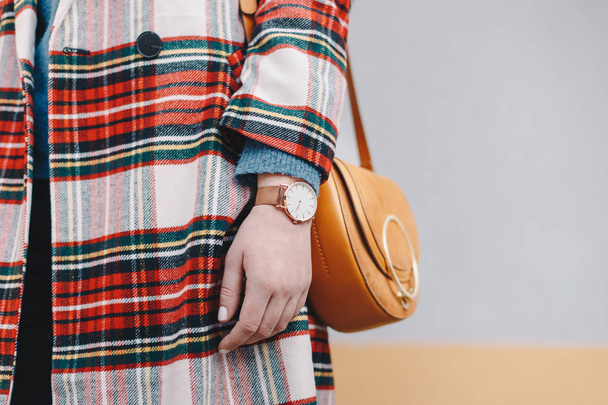 Zavřete detail stylová mladá žena nosí kabát vzor tartanu a náramkové hodinky podržíte ozdobný žluté pytle před vícebarevné pozadí. Prostor pro kopírování, text, reklama. - Fotografie, Obrázek