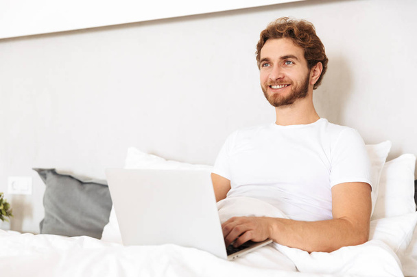 Image of a handsome young bearded man in bed at home using laptop computer. - Valokuva, kuva