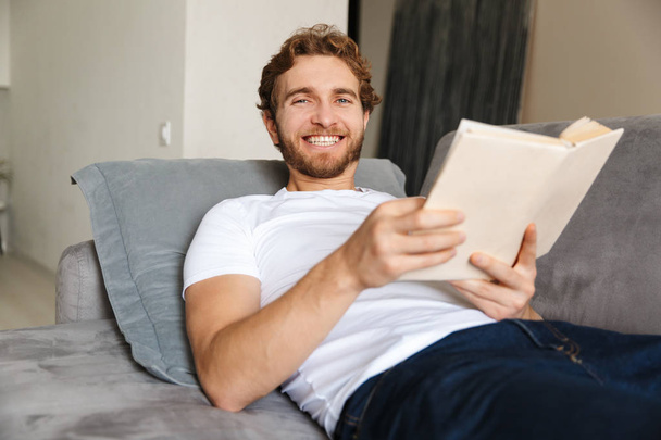 Image of a handsome young bearded man on sofa at home reading book. - Foto, Imagen