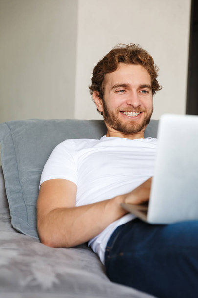 Image of a handsome young bearded man on sofa at home using laptop computer. - Photo, image