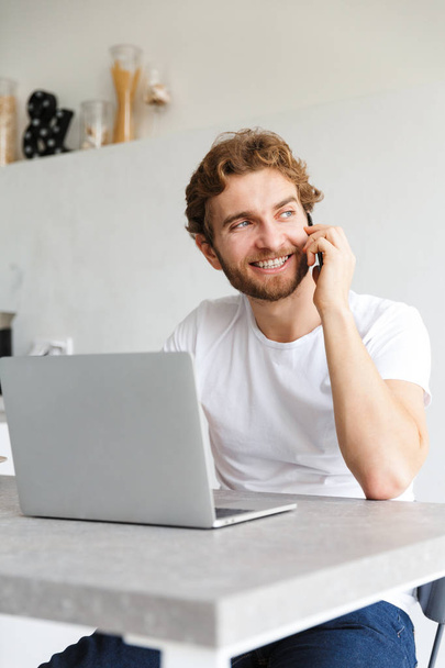 Photo of a happy young bearded man at the table at home talking using laptop computer talking by phone. - Foto, Imagem