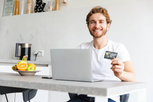 Фото щасливого молодого бородатого чоловіка за столом вдома розмовляють за допомогою ноутбука, який тримає кредитну картку
. - Фото, зображення