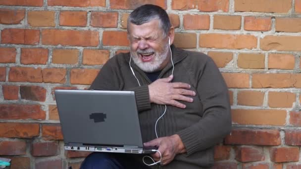 Brodaty starszy człowiek patrząc na ekran notebooka i śmiejąc się siedząc odkryty przed mur z cegły - Materiał filmowy, wideo